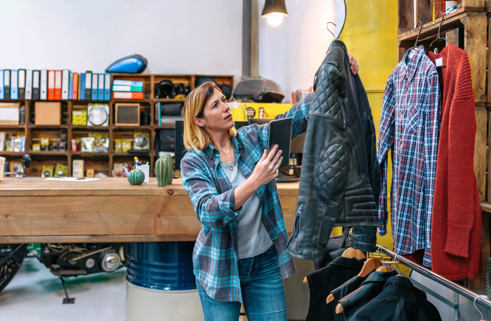 Femme tenant une tablette numérique tout en vérifiant un modèle de veste en cuir noir dans un magasin de vêtements d’occasion. Jeune femme travaillant dans une boutique. Concept d’économie circulaire et de shopping durable.